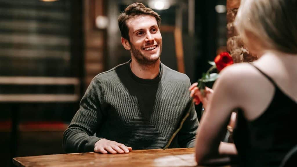 A man smiling at a woman holding a red rose across the table.