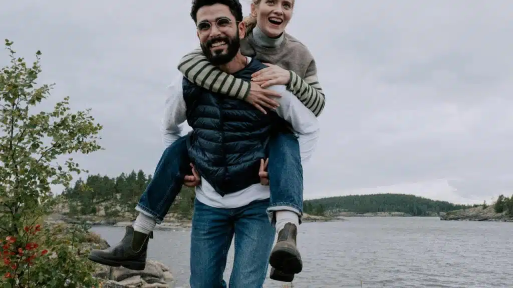 A man giving a piggyback ride to a woman by a lake.
