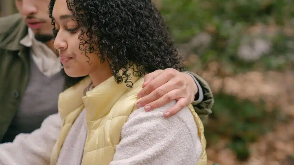 A man comforting a woman with his arm around her shoulder outdoors.