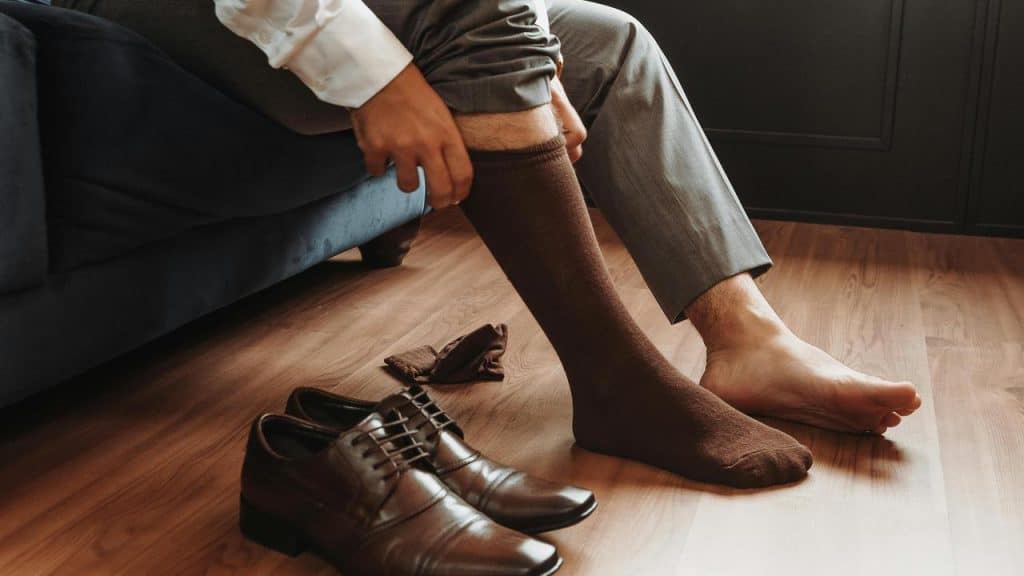 A man sitting on a couch putting on brown socks beside his dress shoes.
