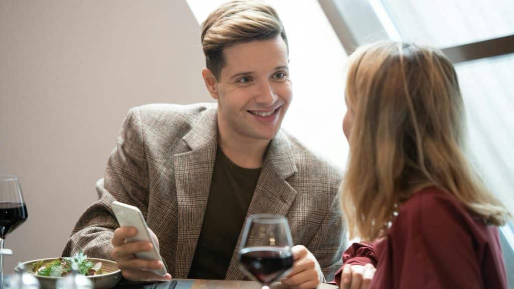 A man smiling at a woman while holding a phone at a restaurant table.