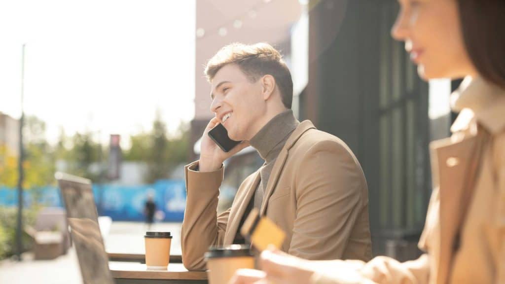 A man smiling while talking on the phone at an outdoor cafΓ©.