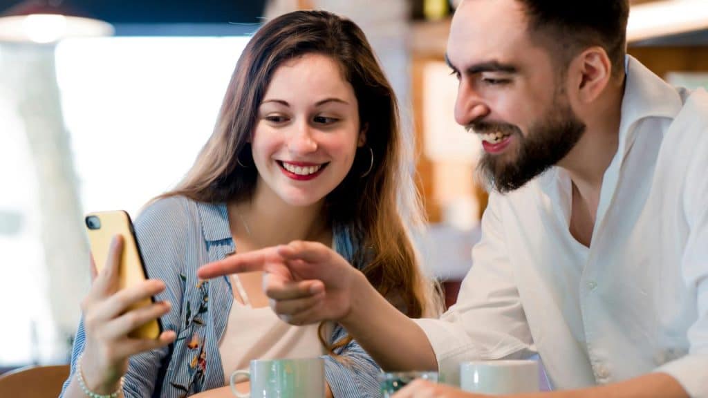 A smiling couple looking at a phone together in a cafΓ©.