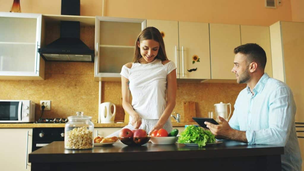 A woman preparing food in the kitchen while a man watches with a tablet.