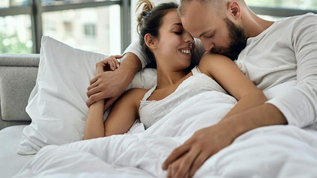 A couple cuddling in bed smiling at each other.