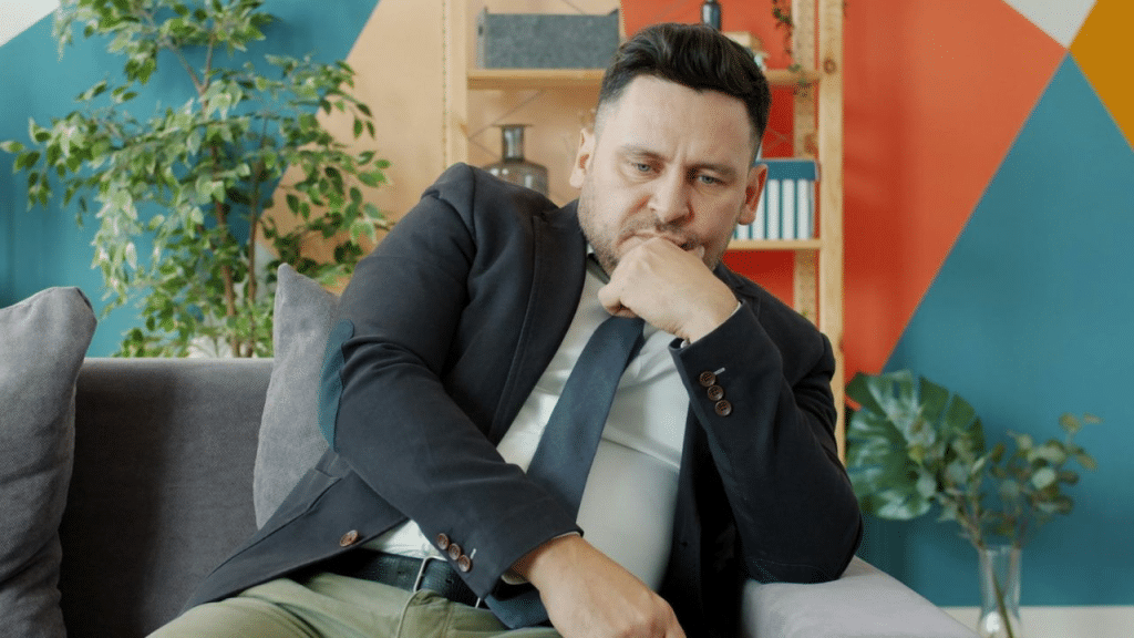 A man in a suit and tie sits on a couch, looking thoughtful and concerned.