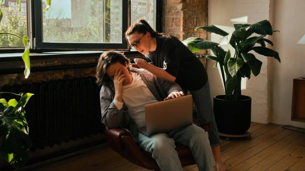 A woman leans over to talk to a man sitting in a chair with a laptop and his hand on his face.