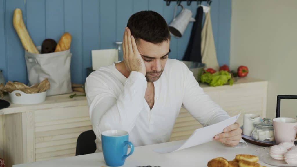 Man in a white shirt sits at a table, reading paper with his hand on his head.