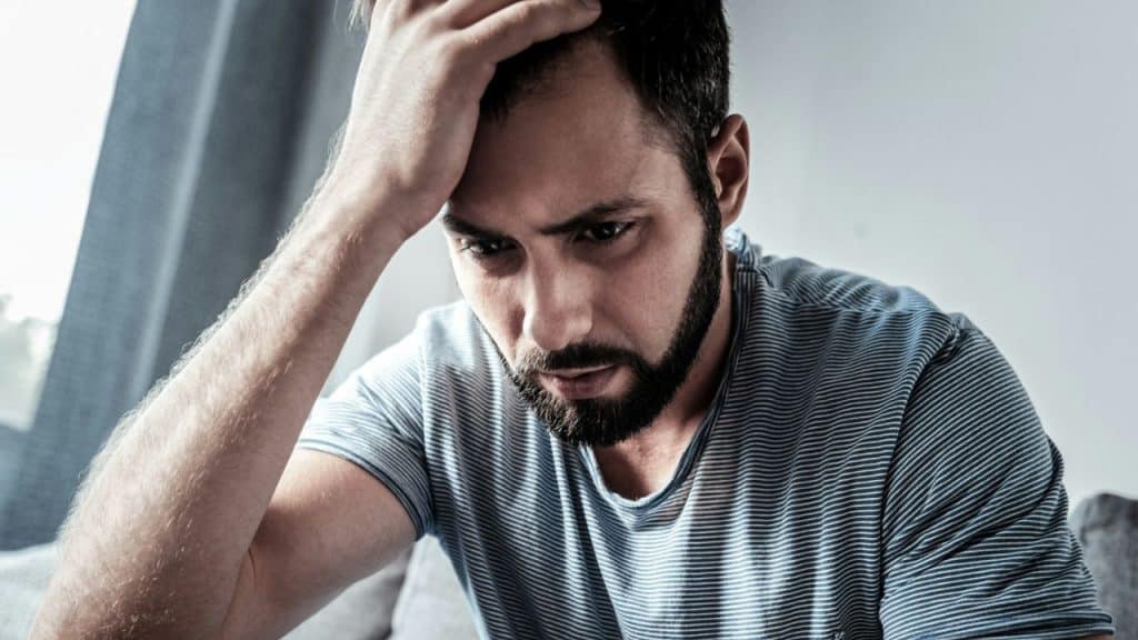 Close-up of a bearded man with a distressed expression, holding his hand to his forehead.