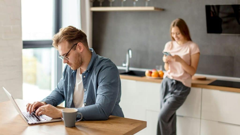 A man works on a laptop at a kitchen table while a woman uses a phone in the background.