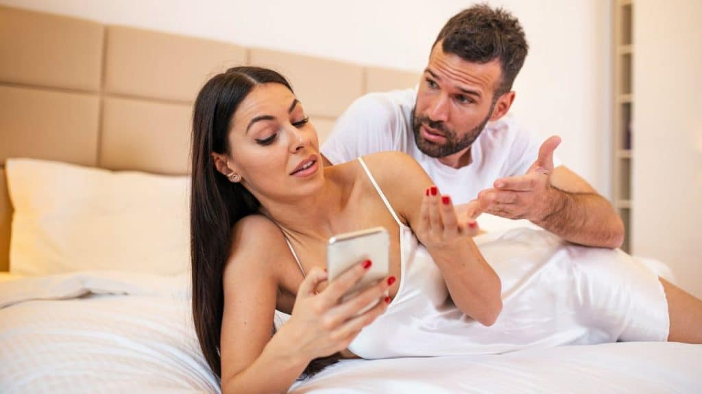A woman lying in bed looks at a phone while a man sitting behind her argues with a confused expression.