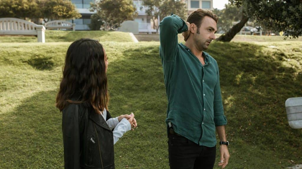 A man standing on the grass with his hand on his neck while a woman looks at him.