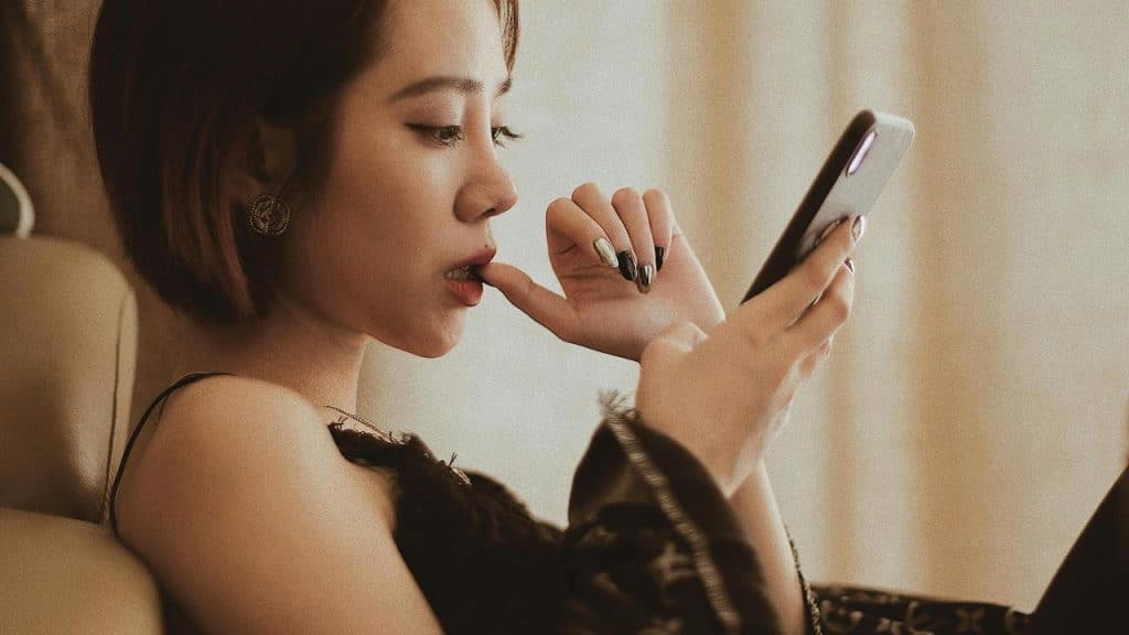 A woman sitting and looking at her phone while biting her finger.