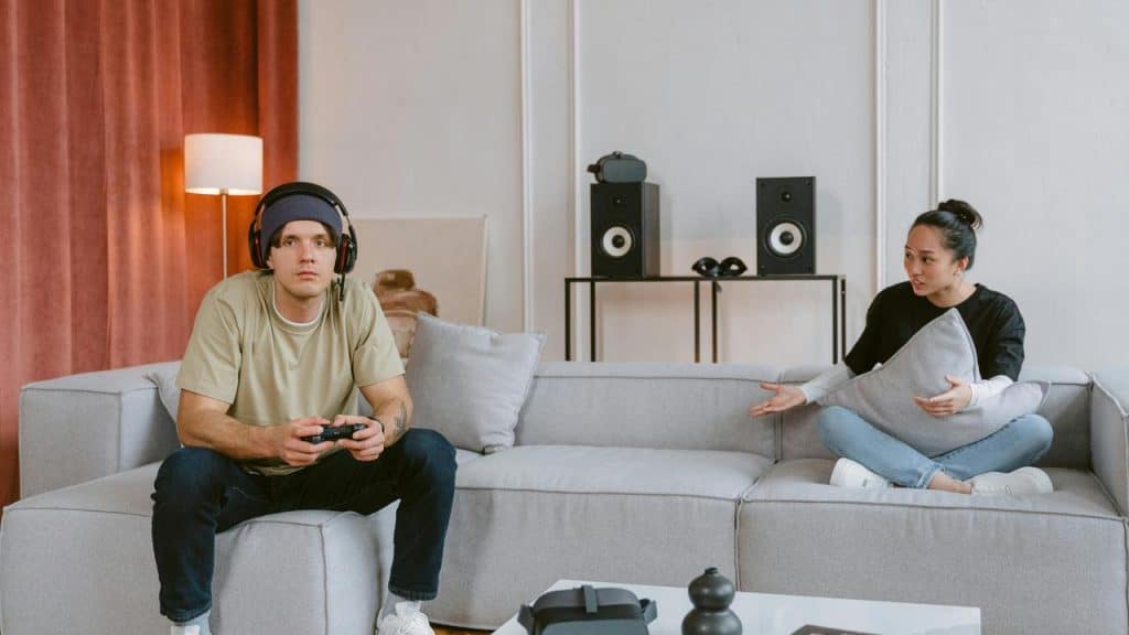 A man gaming with headphones while a woman gestures toward him on the couch.