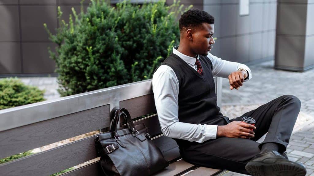 A man in business attire sitting on a bench checking his watch.