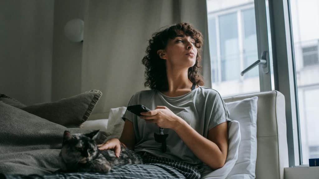A woman sitting on a bed with a cat, holding a TV remote and looking out the window.