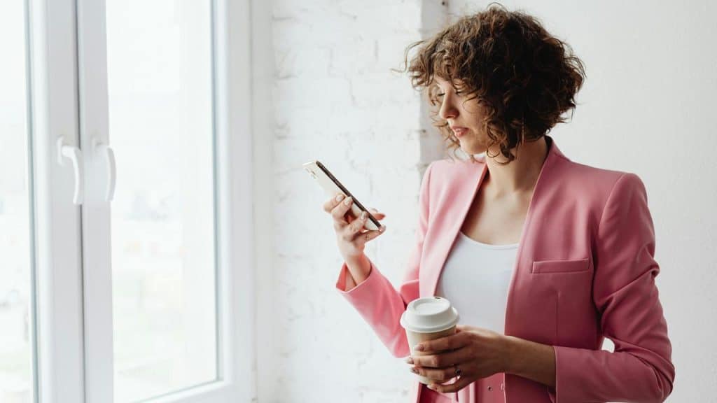 A woman in a pink blazer holding a coffee cup and looking at her phone.