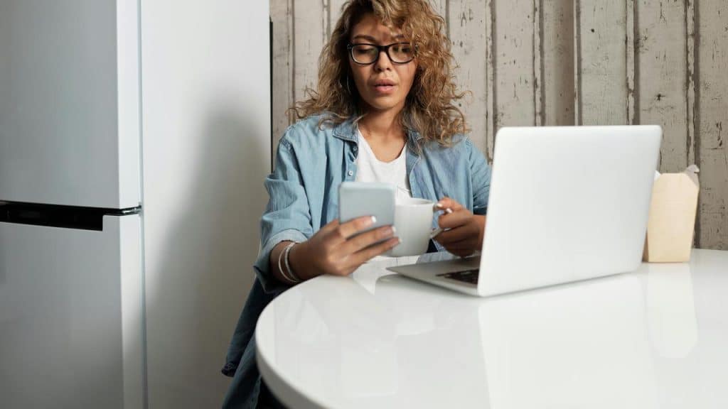 A woman sitting at a table with a laptop, holding a phone and a coffee cup.