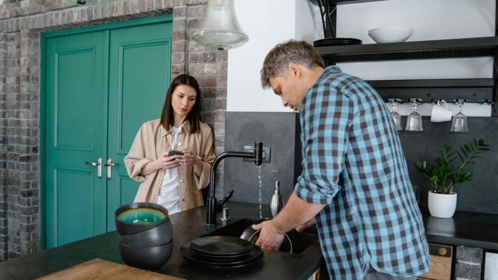 A man washing dishes while a woman stands nearby looking at her phone.