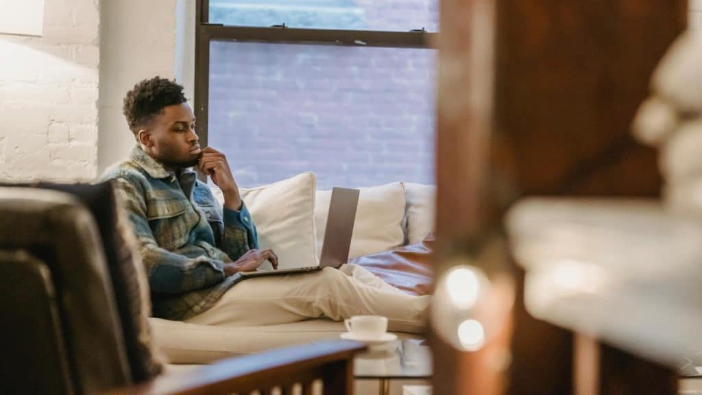 A man sitting on a couch working on a laptop with a thoughtful expression.
