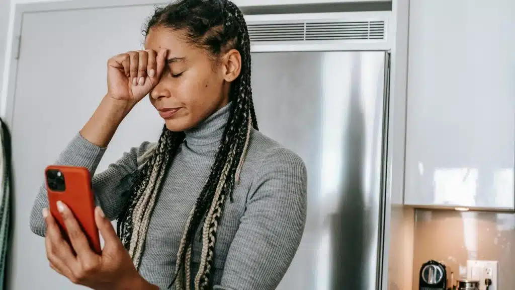 A woman holding her phone and rubbing her forehead with a frustrated expression.