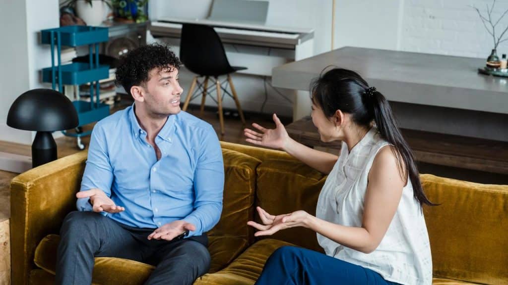 A man and a woman sitting on a couch arguing with raised hands.