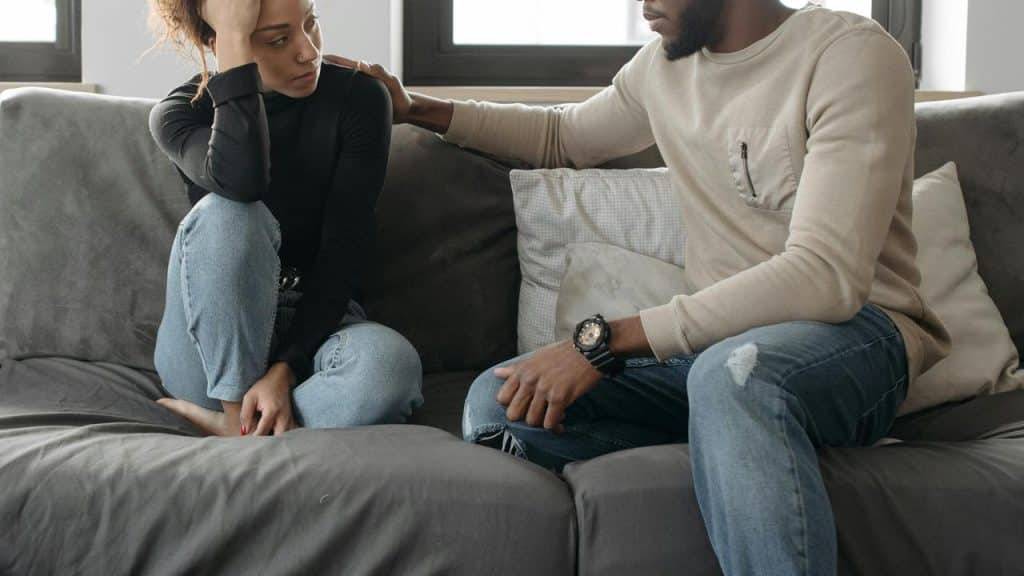 A man comforting a woman sitting on a couch with a sad expression.
