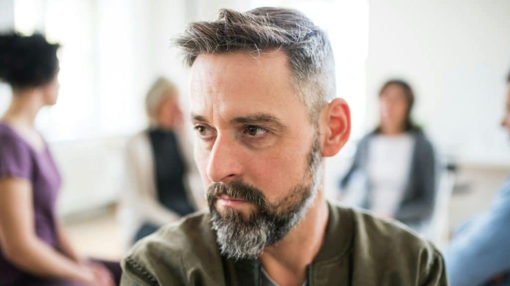 Middle-aged man with a beard and gray hair looking thoughtful in a group setting.