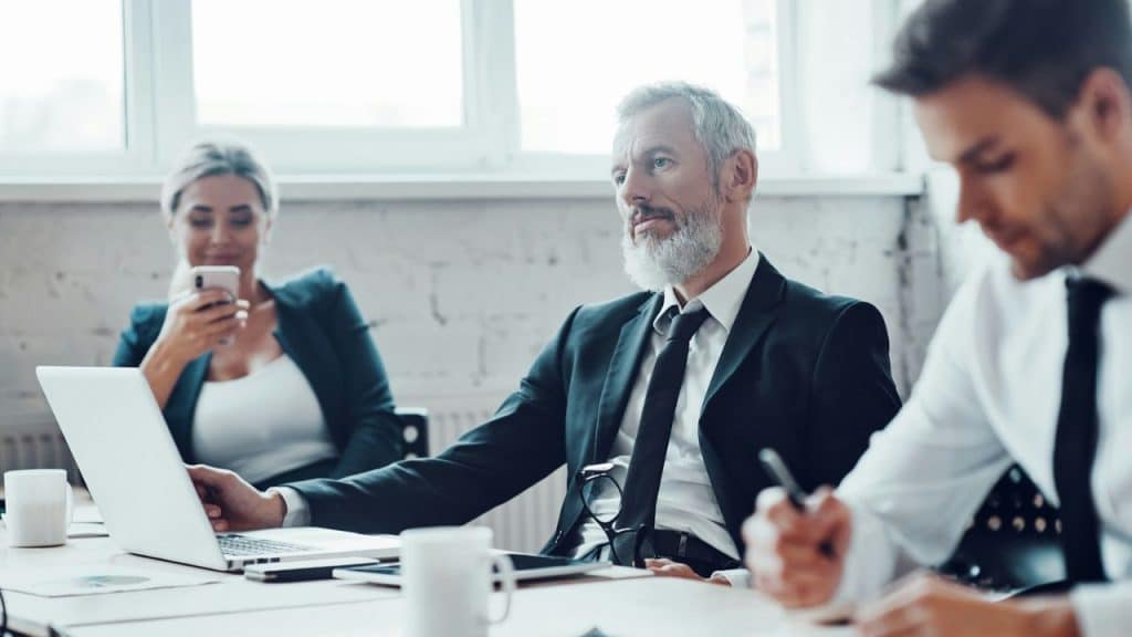 Senior executive in a suit and beard sitting at a conference table with colleagues.