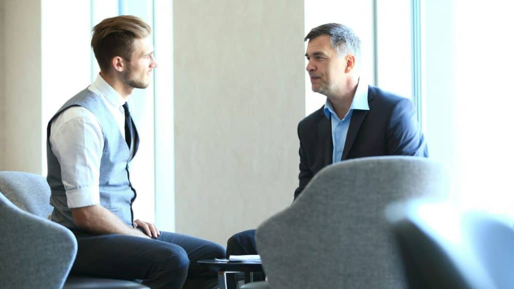 Two businessmen in formal attire talking while sitting in chairs by a bright window.