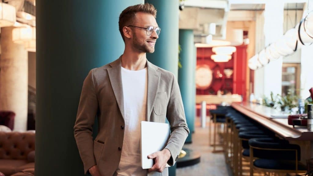 Smiling man in a beige blazer and glasses holding a laptop in a modern bar or lounge.