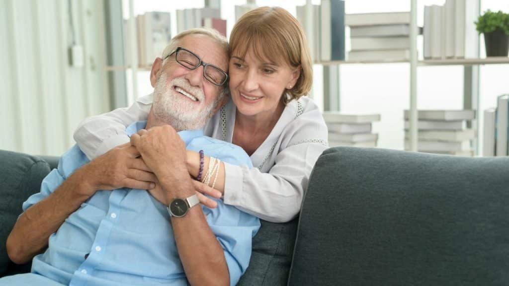 An older woman is embracing a smiling older man on a couch in a brightly lit room.