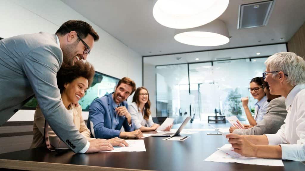 Smiling colleagues discussing papers around a long black table in a bright, modern conference room.