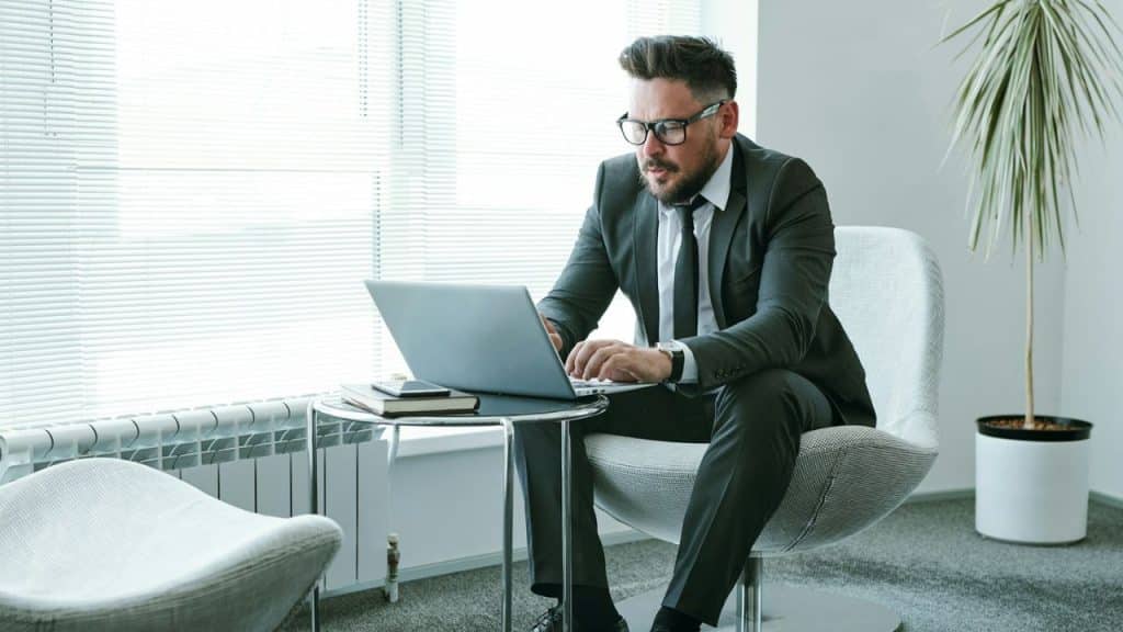 Man in a dark suit and glasses working intently on a laptop next to a window with blinds.