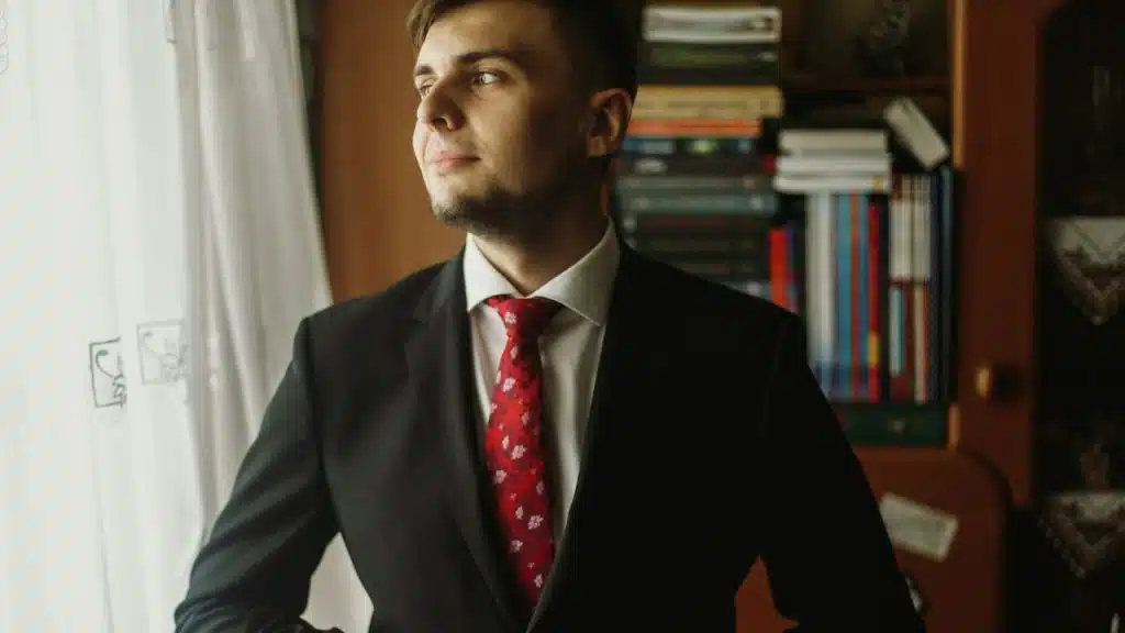 Man in a black suit and red floral tie standing by a window and a bookshelf.
