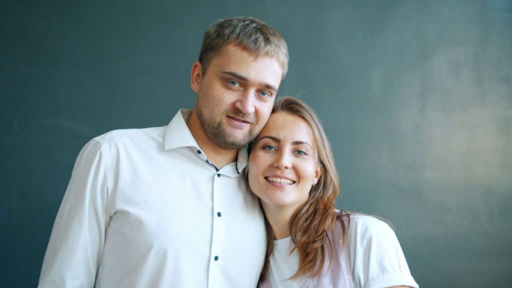 Smiling couple, a man in a white shirt and a woman leaning on his shoulder, against a dark background.