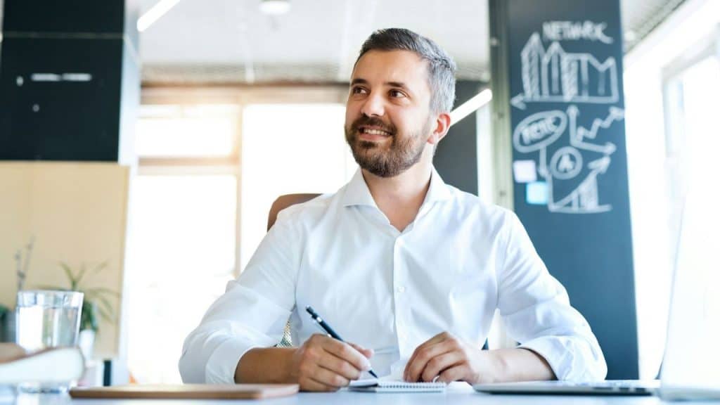 Smiling, bearded man in a white shirt writing at a desk with a chalkboard drawing behind him.