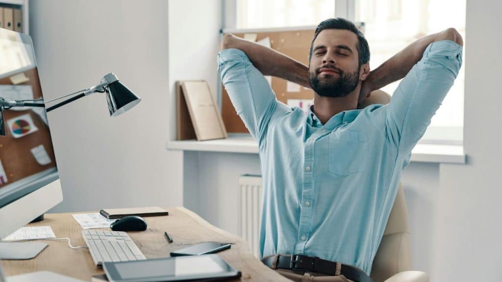 Man in a light blue shirt leaning back in his office chair with hands behind his head and eyes closed.