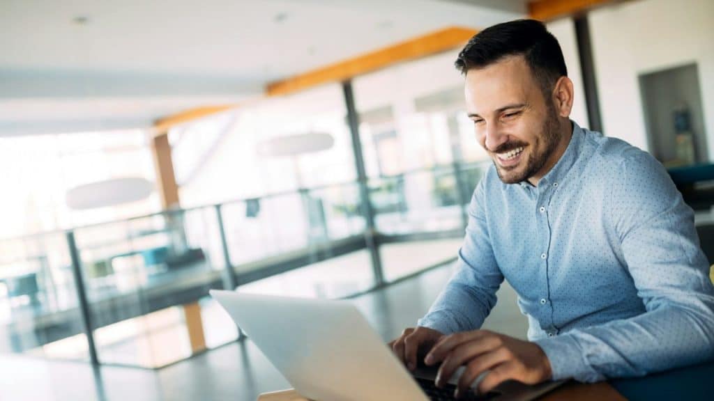 Smiling man in a blue button-down shirt working on a laptop in a bright, modern office space.