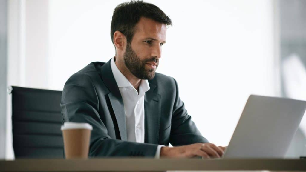 Man in a suit working on a laptop at a desk with a coffee cup in a brightly lit office.