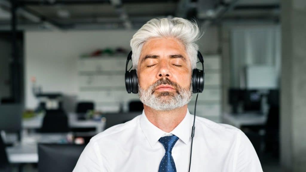Older man in a white shirt and tie with his eyes closed, wearing over-ear headphones in an office setting.