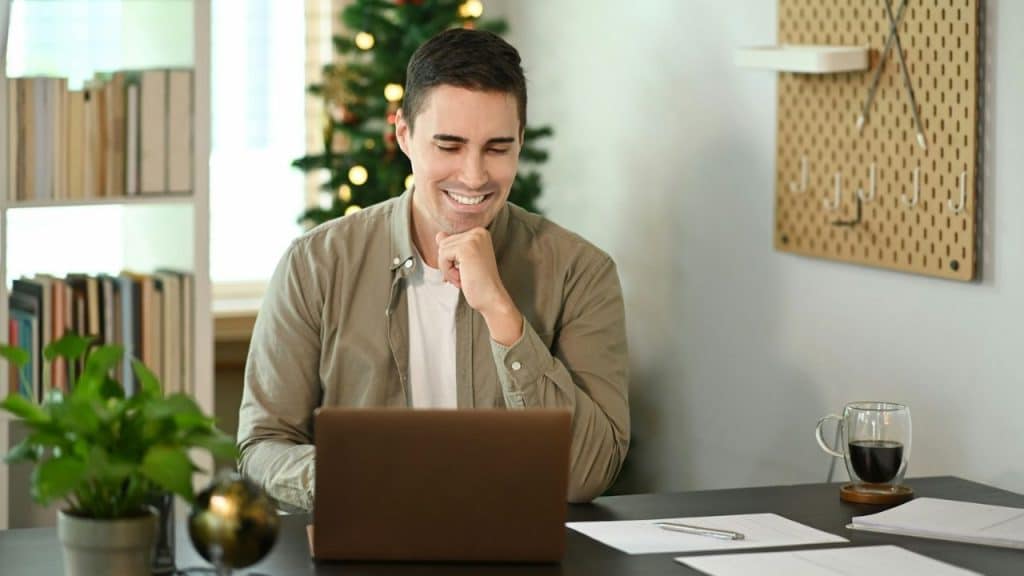 Smiling man working on a laptop at a desk with a Christmas tree visible in the background.