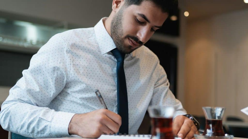 Man in a white patterned shirt and blue tie writing in a notebook at a table with small glasses of tea.