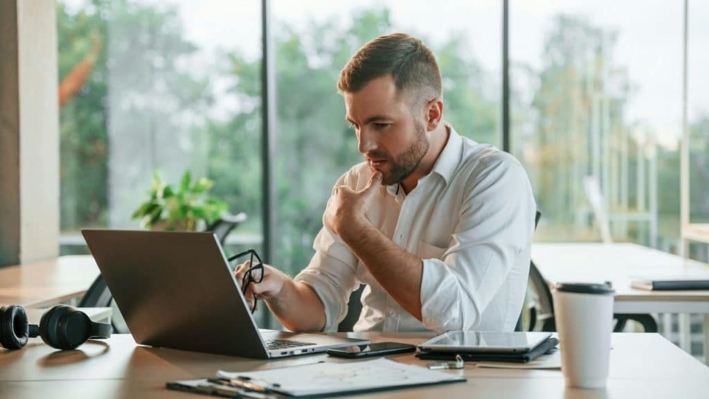 Man in a white shirt looking intently at his laptop screen with his hand on his chin in a bright office.