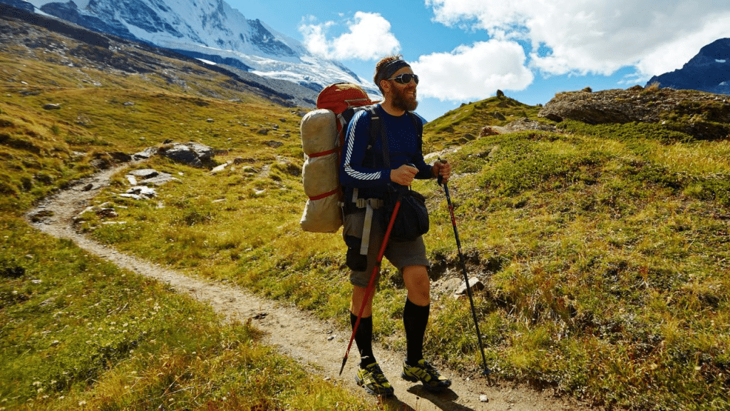 A smiling bearded man with a large backpack hikes on a sunny mountain trail.