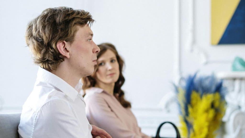 A man sitting in profile while a woman looks at him in the background.