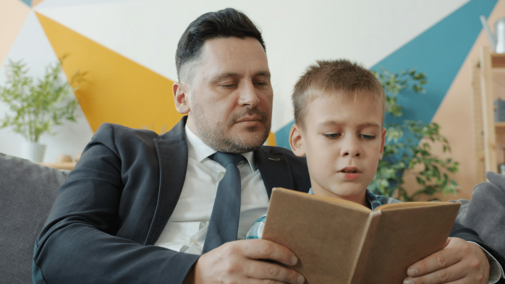 Father in suit reading a book with his young son on a couch indoors.
