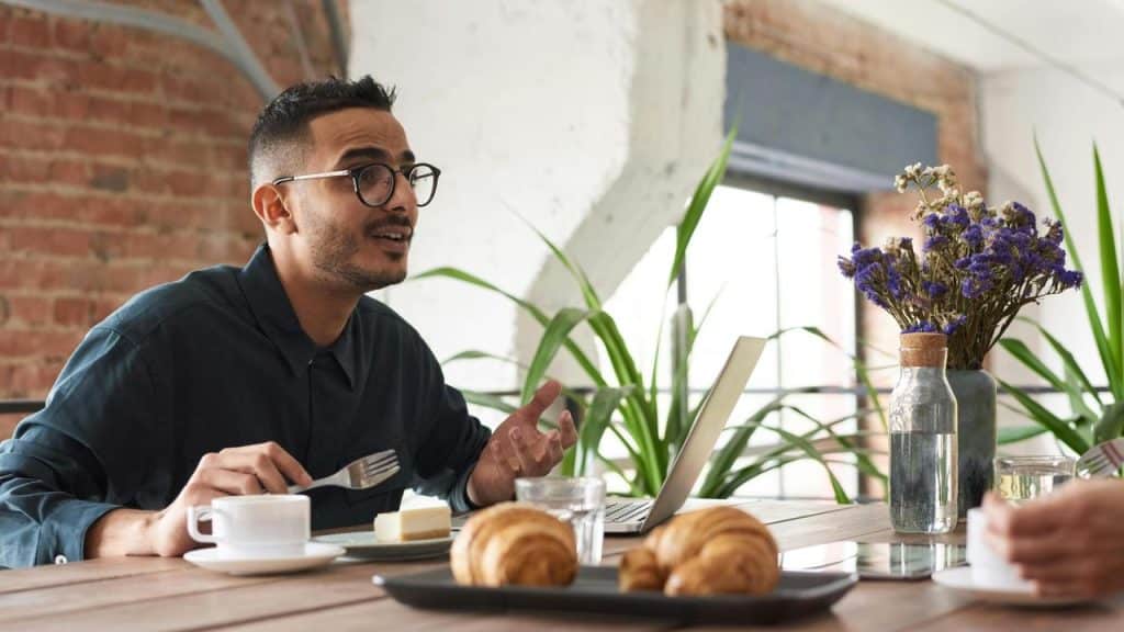 A man talking while having coffee and pastries at a table with a laptop.