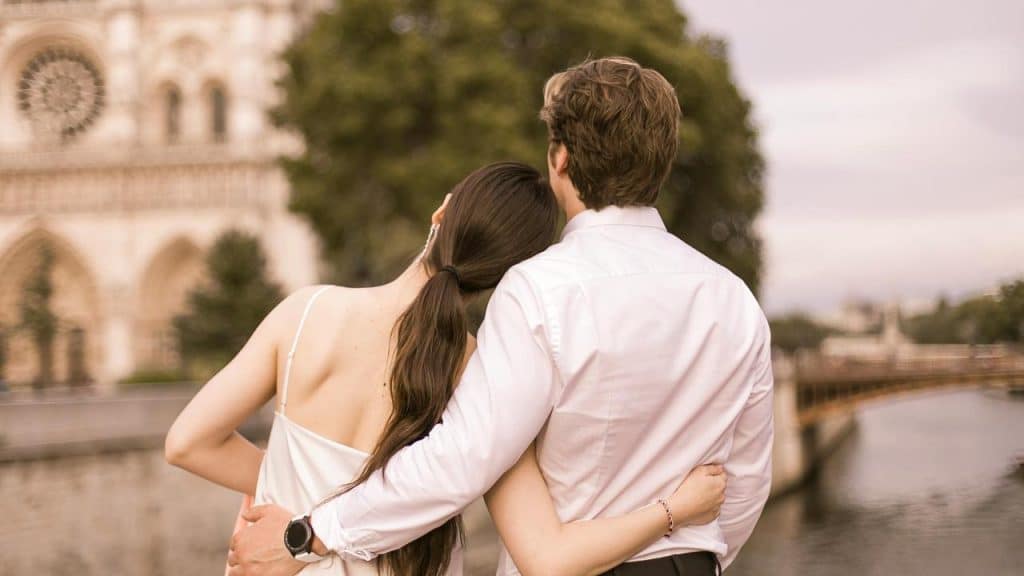 A couple standing by the water with the woman resting her head on the man’s shoulder.