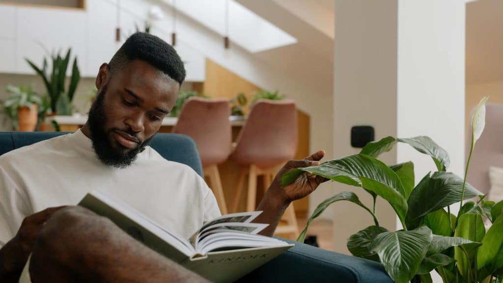 A man sitting on a chair reading a book next to a plant.